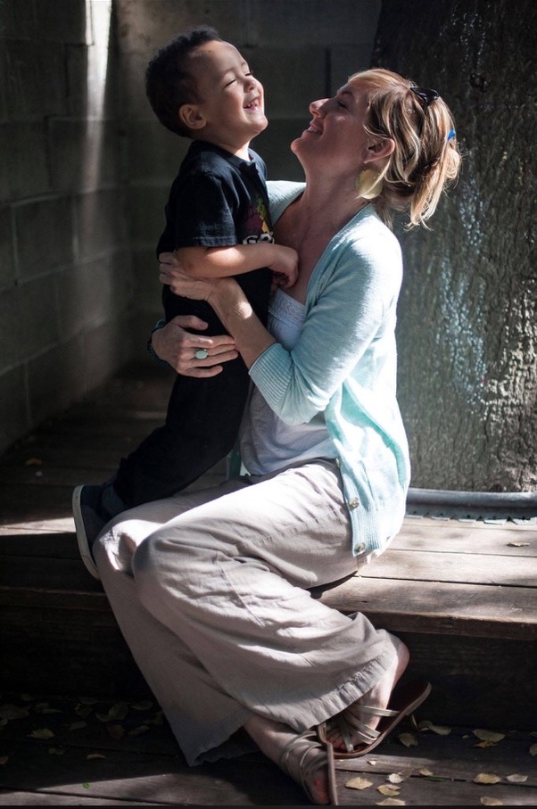 Aimee Gandy smiling with a young child in her arms, both laughing in soft afternoon light