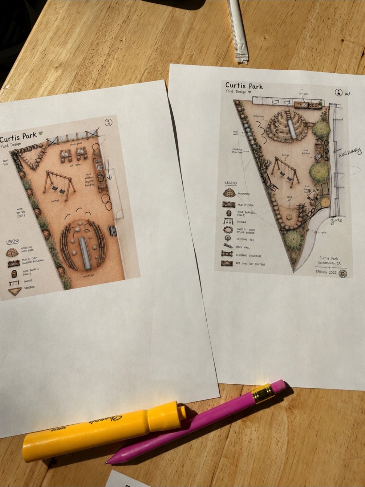 Two printed play yard sketches with handwritten notes, side by side on a wooden café table with a marker and pencil